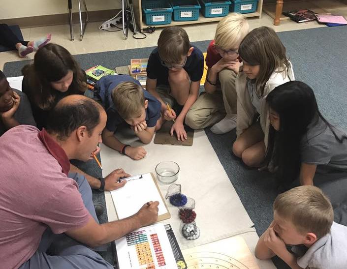 Teacher sits on floor with students guiding them in a math lesson