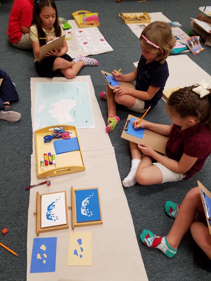 Students sitting on the floor around a Montessori work, taking notes